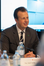 Person in dunklem Anzug spricht auf einem Podium, Konferenzsituation.