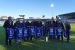 Bildbeschreibung: Eine Gruppe von Menschen in sportlicher Kleidung auf einem Fußballfeld, halten blaue Trikots mit Nummern hoch.