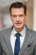 Portrait einer männlichen Person mit dunklem Haar, Anzug, Hemd und blauer Krawatte, die direkt in die Kamera schaut