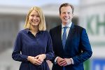 Bildbeschreibung: Zwei lächelnde Personen im professionellen Outfit stehen nebeneinander.