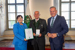 Drei Personen im feierlichen Rahmen. Zwei Personen überreichen eine Urkunde an eine dritte Person.