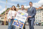 Drei Personen präsentieren ein Plakat mit Kindergesichtern und dem Aufdruck "Landesfinale Steiermark on Tour" vor einem Stadtbild.