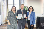 Drei Personen stehen lächelnd vor einer Wand. In der Mitte hält eine Person eine Urkunde.
