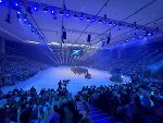 Bildbeschreibung: Großer Saal mit Publikum und Musikern und Militärfahrzeugen auf der Bühne.
