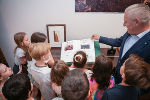 Gruppe Kinder lauscht interessiert einer Person, die ein Buch vorliest. Im Hintergrund ein Raum mit Gemälden.