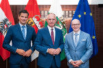 Drei Personen in Anzügen stehen vor Flaggen von Österreich, Steiermark und der EU.