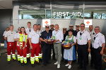 Gruppe in Uniformen vor einem Gebäude mit dem Aufdruck 'FIRST AID'. Eine Person überreicht eine gefüllte Körbchen.