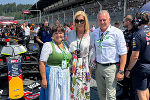 Drei lächelnde Menschen stehen vor Formel-1-Wagen. Im Hintergrund eine Rennstrecke mit Zuschauern.
