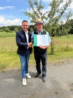 Bildbeschreibung: Zwei Menschen im Gespräch und Händedruck bei einer Preisverleihung in ländlicher Umgebung.