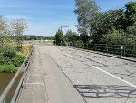 Bildbeschreibung: Blick auf eine asphaltierte Brücke mit Geländer. Im Hintergrund sind Bäume und ein blauer Himmel zu sehen.