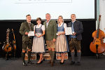 Gruppe in Tracht auf Bühne, hält Auszeichnungen. Im Hintergrund Musikinstrumente und eine Leinwand.