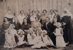 Bildbeschreibung: Historisches Gruppenfoto von Personen, teilweise in Ordensgewändern und Schürzen, vor einem Holzhintergrund.
