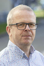 Bildbeschreibung: Porträt eines Mannes mit Brille und gemustertem Hemd vor einem unscharfen Hintergrund.