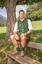 Person in traditioneller Tracht sitzt auf einer Holzbank im Grünen.